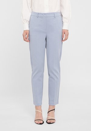 Femme portant un pantalon bleu clair sur mesure longueur cheville, une blouse blanche et des sandales à talons hauts noires et beiges à brides fines.