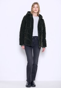 Giacca a vento verde scuro con cappuccio, caratterizzata da un design trapuntato e lacci regolabili. Abbinata a jeans grigi e stivali neri.