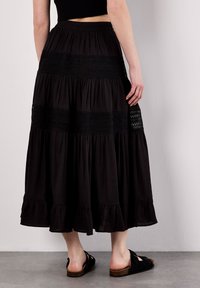 Gonna maxi nera con design a strati, caratterizzata da dettagli in pizzo e orlo arricciato. Indossata con sandali neri casual, che mettono in risalto la fluidità del tessuto.