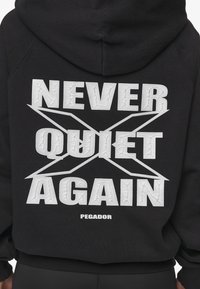 Sudadera negra con gran texto blanco que dice "NUNCA MÁS CALLADO" y una etiqueta de marca "PEGADOR" en la parte trasera. Material de algodón.