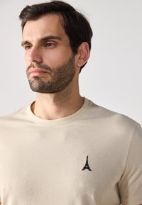 Beige bomull t-skjorte med en liten svart brodert Eiffel-tårn-design på venstre brystområde. Enkel design med rund hals.