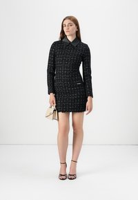 Schwarzes Kleid aus strukturiertem Stoff mit einem kontrastierenden schwarzen Kragen. Verfügt über ein figurbetontes Design, einen fransigen Saum und zwei aufgesetzte Taschen. Das Modell hält eine kleine Tasche.