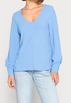 Femme portant un chemisier bleu clair à manches longues avec col en V et poignets boutonnés ainsi qu'un jean délavé clair, debout devant un fond uni.