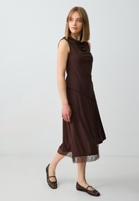 Bruine mouwloze jurk met gedrapeerde halslijn en kantdetail aan de zoom, gecombineerd met zwarte geweven flats. Glad fabric met een zachte textuur.