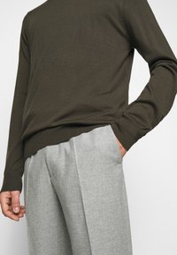 Uomo che indossa un maglione a maniche lunghe verde oliva scuro con una mano nella tasca di pantaloni di lana grigio chiaro, in piedi contro uno sfondo semplice.