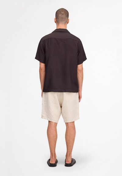 Homme de dos portant une chemise noire à manches courtes, un short beige à hauteur des genoux et des sandales noires sans brides sur fond blanc.