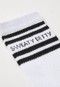 Calcetín de algodón blanco con textura de toalla, que presenta dos rayas negras y la inscripción "SWEATY BETTY" en letras negras.