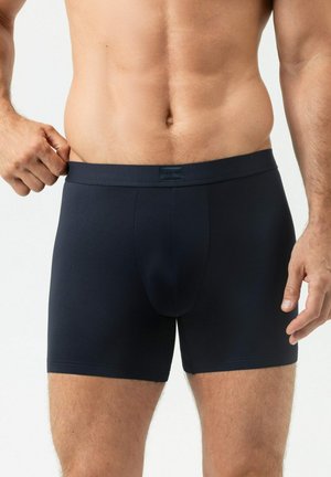 Homme portant un boxer bleu marine foncé, tirant légèrement la ceinture vers l'extérieur, torse et cuisses supérieures visibles sur un fond blanc.
