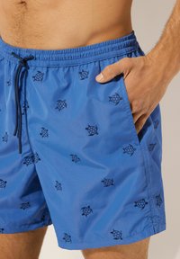 Costume da bagno blu realizzato in tessuto leggero, con un motivo di piccole tartarughe nere, cintura elastica e tasca laterale.