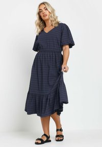 Mekko, jossa on tummansininen väri, lyhyet hihat, ruudullinen kuvio, V-pääntie, elastinen vyötärö ja kerroksellinen röyhelöhko. Yhdistetty mustien sandaalien kanssa.