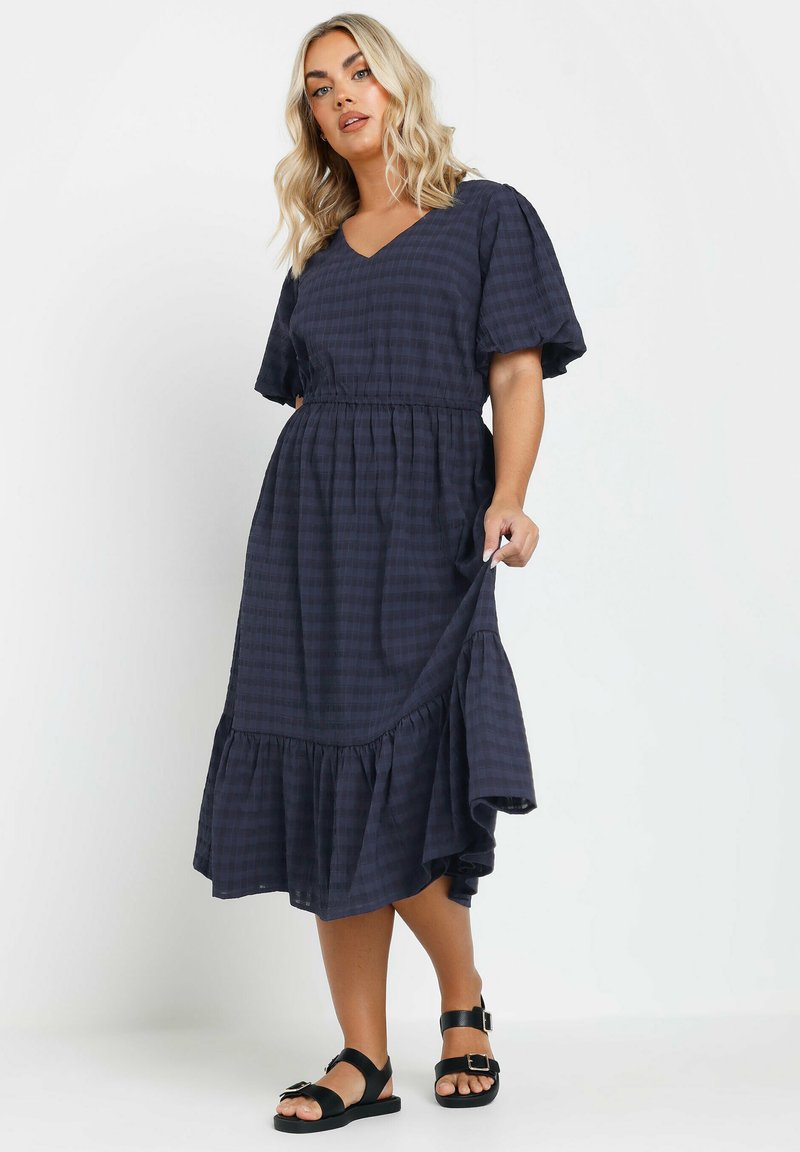 Mekko, jossa on tummansininen väri, lyhyet hihat, ruudullinen kuvio, V-pääntie, elastinen vyötärö ja kerroksellinen röyhelöhko. Yhdistetty mustien sandaalien kanssa.