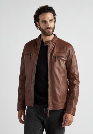 Man met krullend haar, gekleed in een brown leren jas over een zwart shirt en donkere spijkerbroek, kijkt iets naar rechts.