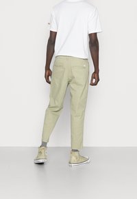 Man med beige kapade byxor, grå strumpor, beige Converse-sneakers och en vit T-shirt, stående med ryggen mot en enkel bakgrund.