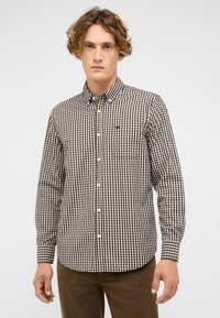 Mann trägt ein braun-weiß kariertes Hemd mit Knöpfen und Brusttasche, kombiniert mit braunen Hosen, steht vor einem einfarbigen hellen Hintergrund.