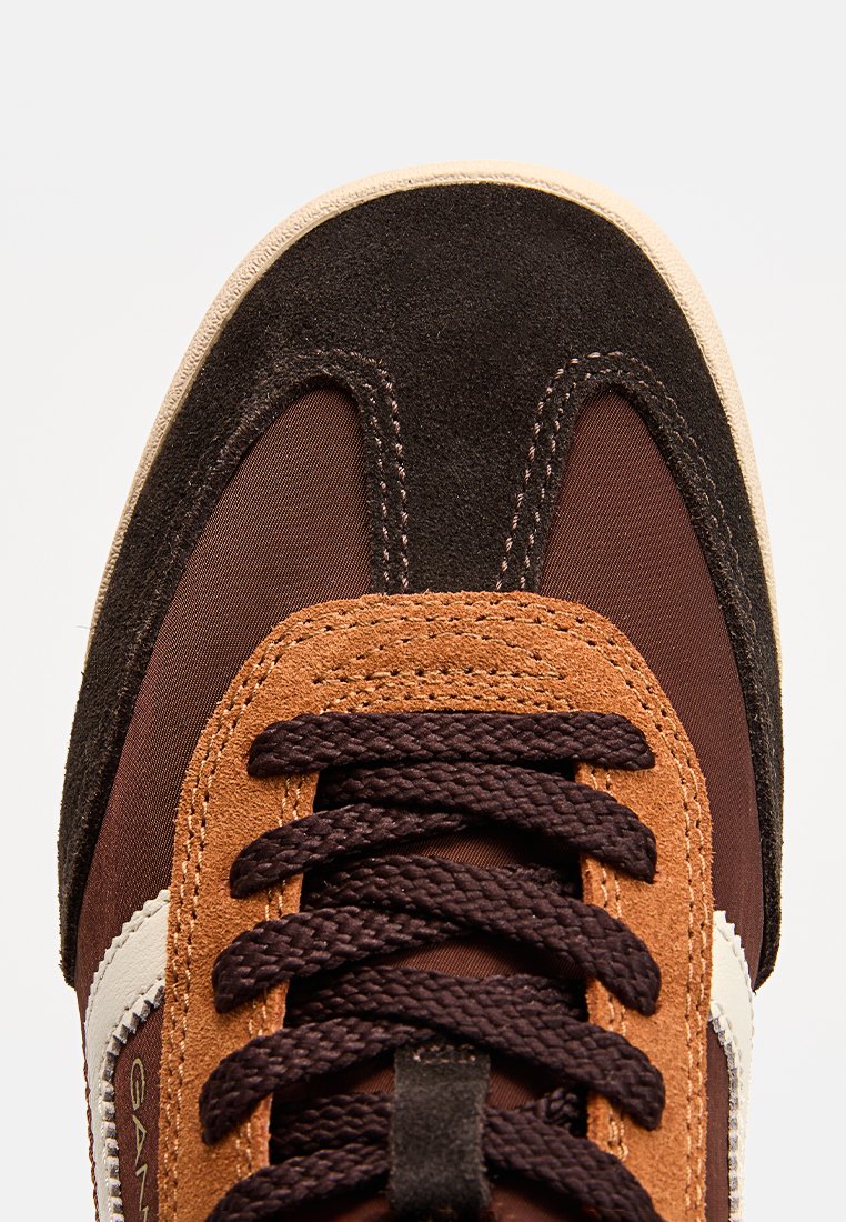 Braune und beige Sneakers mit einem Obermaterial aus Wildleder und Textil, ausgestattet mit dunkelbraunen Schnürsenkeln, einem kontrastierenden weißen Seitenstreifen und einer abgerundeten Zehenpartie.