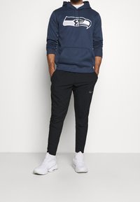 Felpa blu navy con logo dei Seahawks, pantaloni neri a gamba stretta e scarpe da ginnastica bianche. La felpa presenta una tasca frontale e un cappuccio con cordino.