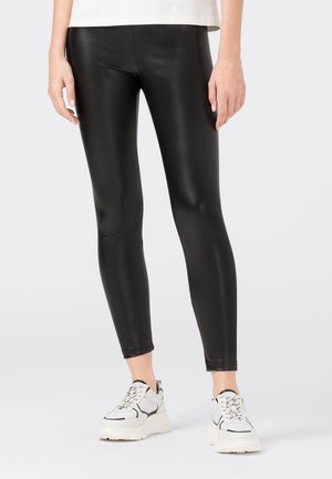 Leggings noirs taille haute en matériau extensible et brillant. Présentent une coupe ajustée et une texture lisse, accompagnés de baskets blanches.