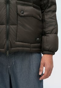 Pufferjakke i mørk olivengrønn, med glidelås foran, to lommer med knapp på brystet, og elastiske mansjetter; brukt over grå bukser.