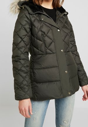 Femme portant une veste d'hiver matelassée vert foncé avec capuche en fourrure et un jean bleu clair déchiré, regardant vers l'avant avec les mains détendues le long du corps.