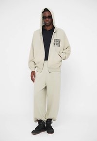 Lichtbeige hoodie met zakken en tekstontwerp, gecombineerd met bijpassende beige broek. Model draagt zwarte sneakers en een zonnebril.