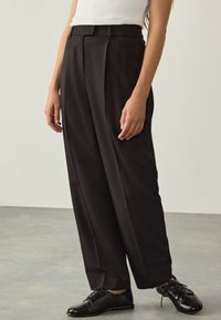 Pantalon noir taille haute à jambes larges, avec des plis sur le devant et une texture lisse, associé à des chaussures noires à lacets.