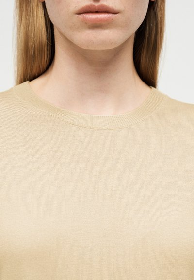 Gros plan sur le cou et la bouche d'une personne portant un pull ras du cou beige, avec des cheveux blonds raides visibles.