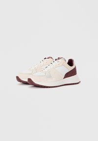Un paio di sneakers basse beige e bordeaux con lacci bianchi e accenti in tessuto testurizzato su uno sfondo bianco uniforme.
