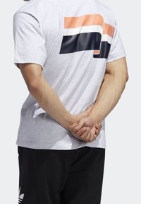 Šedé tričko s krátkým rukávem, které má geometrické logo v oranžové, bílé a námořnické modři na horní části zad, nošeno s černými kalhotami.