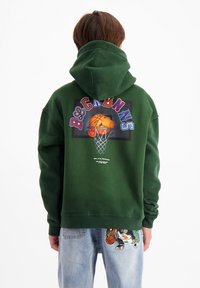 Groene hoodie met een grafisch ontwerp van een basketbal en kleurrijke tekst op de achterkant; zachte stof, losse pasvorm en een grote capuchon.