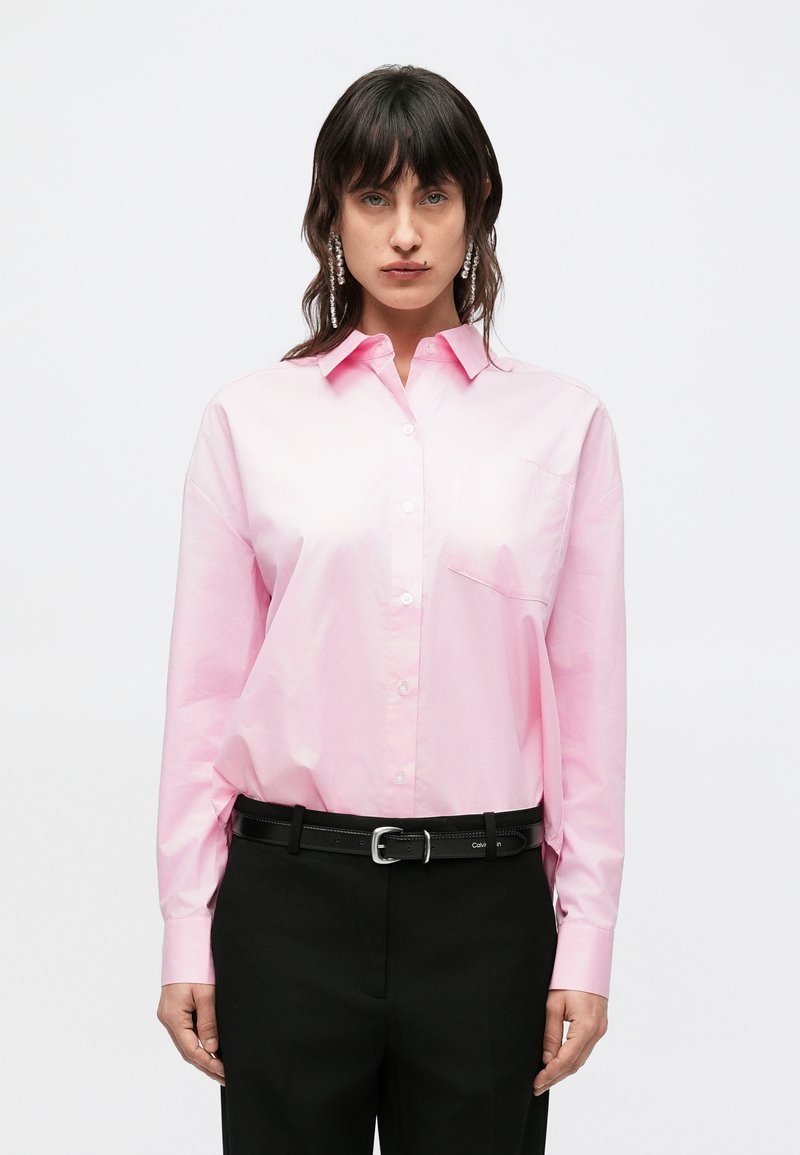 Ružová košeľa na zapínanie z hladkej tkaniny, s golierom a jedným náprsníkom, v kombinácii s čiernymi nohavicami a čiernym opaskom.