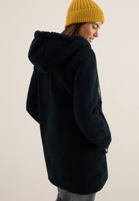 Nainen, jolla on yllään tummansininen fleece-takki hupulla ja sinappikeltainen neulottu pipo, seisoo sivuttain kädet takin taskuissa.