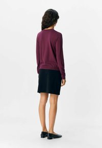Pull en maille bordeaux à manches longues, col rond, associé à une jupe noire. Chaussures plates noires, vues de derrière sur un fond uni.