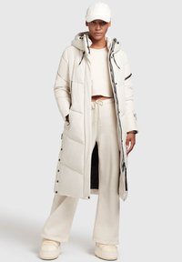 Long manteau matelassé beige avec capuche, coupe oversize, boutons sur le devant et deux poches latérales. Porté par-dessus un haut crème et un pantalon large assorti.