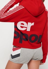 Sudadera con capucha roja y acentos en blanco y marino, con gran texto "Sport". Combinada con pantalones cortos grises que exhiben patrones geométricos.