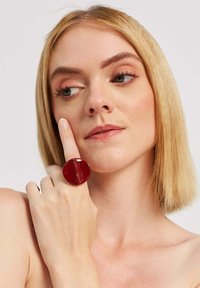 Mirdzošs sarkans gredzens ar apaļu formu, nēsāts uz rādītājpirksta, ar gludu virsmu un sadalītu dizaina detaļu.