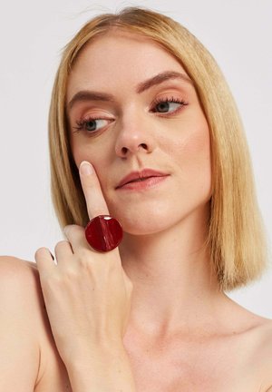 Błyszczący czerwony pierścień o okrągłym kształcie, noszony na palcu wskazującym, z gładką powierzchnią i detalem w postaci rozdzielenia.