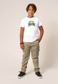 T-shirt blanc avec un motif de voiture verte, associé à un pantalon cargo kaki avec des poches latérales et des baskets noires à semelles blanches.