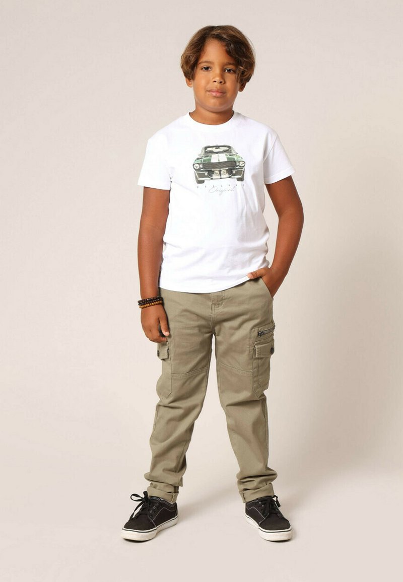 T-shirt blanc avec un motif de voiture verte, associé à un pantalon cargo kaki avec des poches latérales et des baskets noires à semelles blanches.