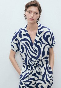 Short-sleeve jumpsuit in cream fabric with navy abstract pattern. Features collar, button front, and tied waist. Pockets on each side.