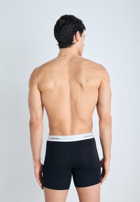 Crne bokserice s sivoj gumi koja nosi logo "Calvin Klein", prikazujući glatku tkaninu i prilagođen dizajn s leđa.