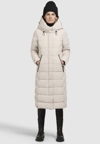 Lang cremet pufferfrakke med hætte, der har et quiltet design, snorede detaljer og lynlåslommer på siden. Et par sorte støvler er synlige.