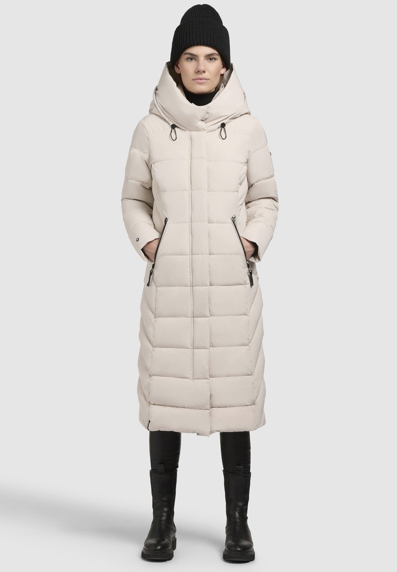 Lang cremet pufferfrakke med hætte, der har et quiltet design, snorede detaljer og lynlåslommer på siden. Et par sorte støvler er synlige.