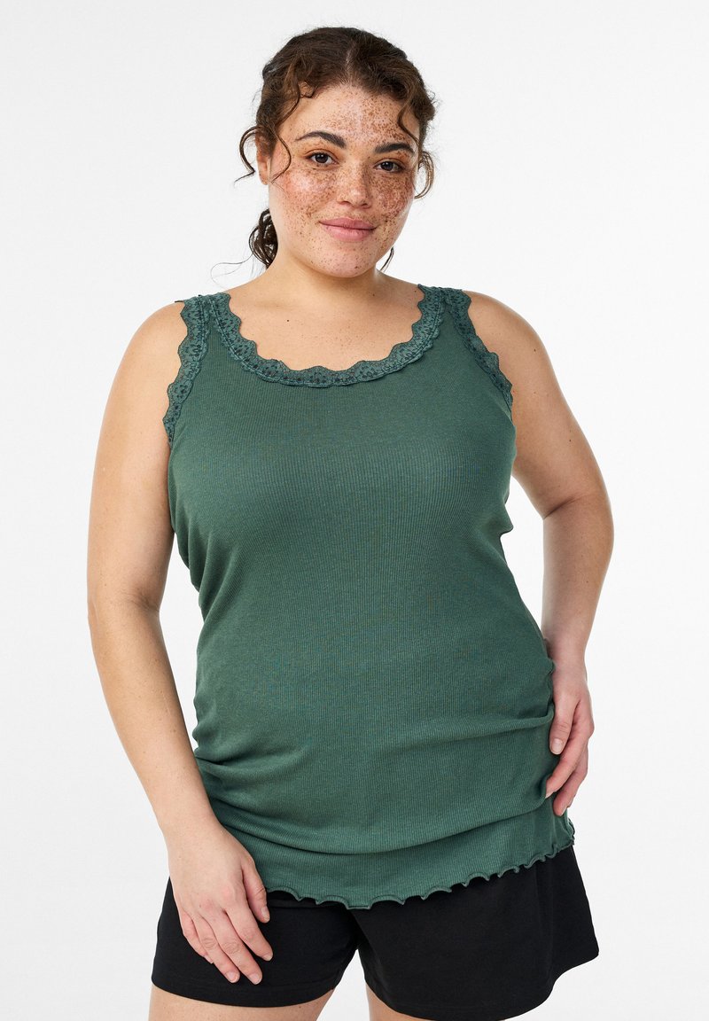 Grünes, geripptes Tanktop mit gewelltem Spitzenbesatz entlang des Ausschnitts und der Träger, mit weicher Textur und lockerem Sitz, kombiniert mit schwarzen Shorts.