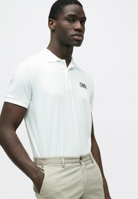 Weißes Poloshirt mit Kragen und schwarzem, gesticktem Logo. Kombiniert mit hellbeigen Hosen, die ein figurbetontes Design und glatte Textur zeigen.