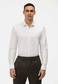 Wit overhemd met lange mouwen en knopen, slim fit, en een strakke kraag, gecombineerd met donkere broek met elastische taille, voor een effen achtergrond.