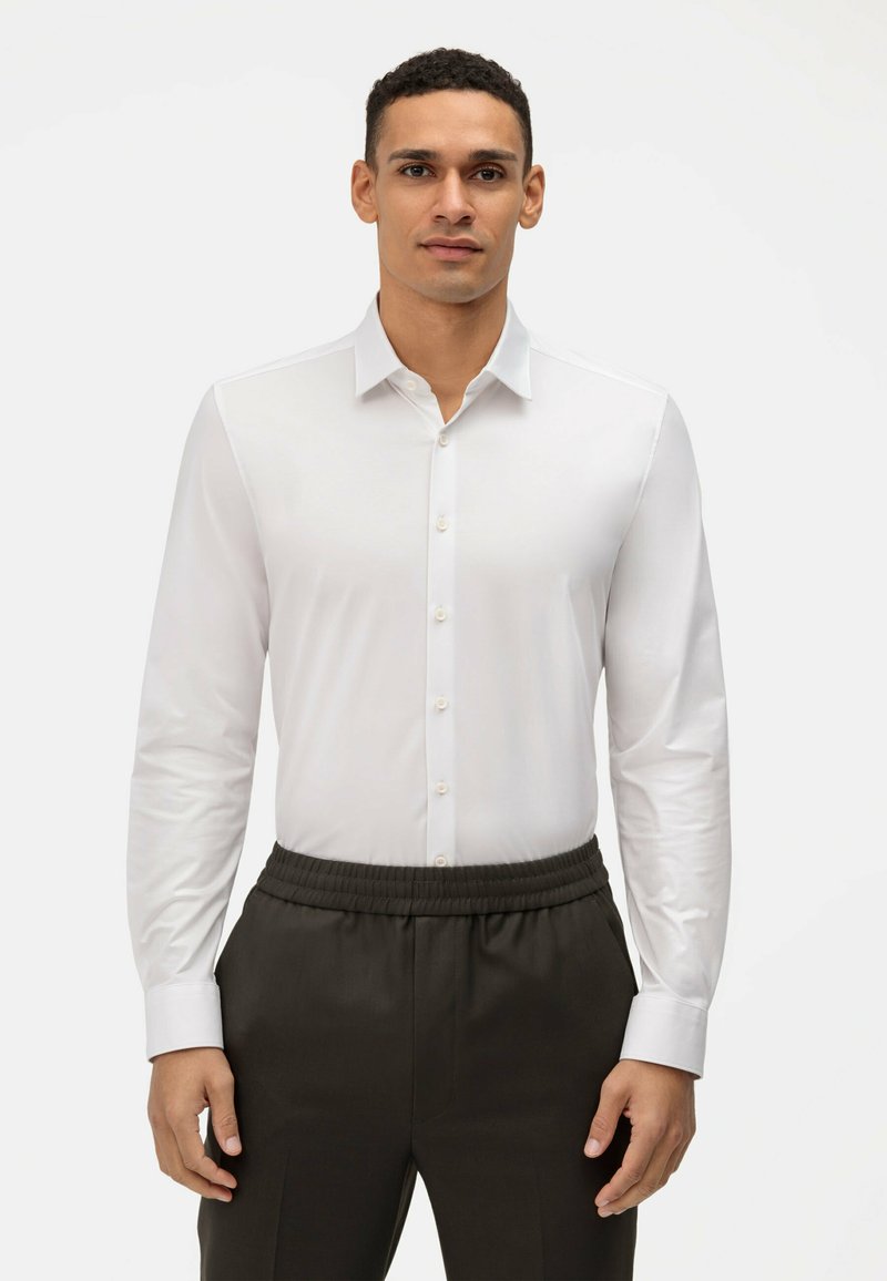 Wit overhemd met lange mouwen en knopen, slim fit, en een strakke kraag, gecombineerd met donkere broek met elastische taille, voor een effen achtergrond.