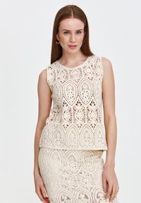 Beżowy bezrękawnik z misternymi wzorami koronkowymi, z motywami kwiatowymi i liściastymi oraz falbaniastym dekoltem. W komplecie ze spódnicą w tym samym stylu.