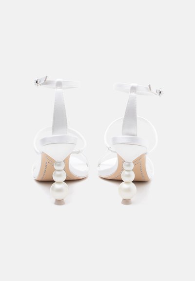 Λευκά σανδάλια με ψηλό τακούνι, λουράκια στον αστράγαλο και τακούνια στοιβαγμένα με στυλ πέρλας, εμφανιζόμενα από πίσω με λευκό φόντο.