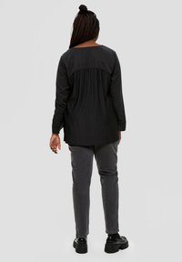 Schwarz Oberteil mit plissiertem Rücken, langen Ärmeln und rundem Ausschnitt; kombiniert mit grauen Slim-Fit-Hosen und schwarzen Plateauschuhen. Neutraler Hintergrund.