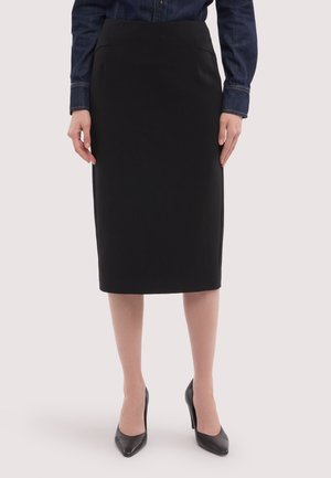 Kvinde iført en sort knælængde blyantskørt, mørk denimskjorte med knapper og sorte spidse høje hæle, stående med hænderne langs siden.